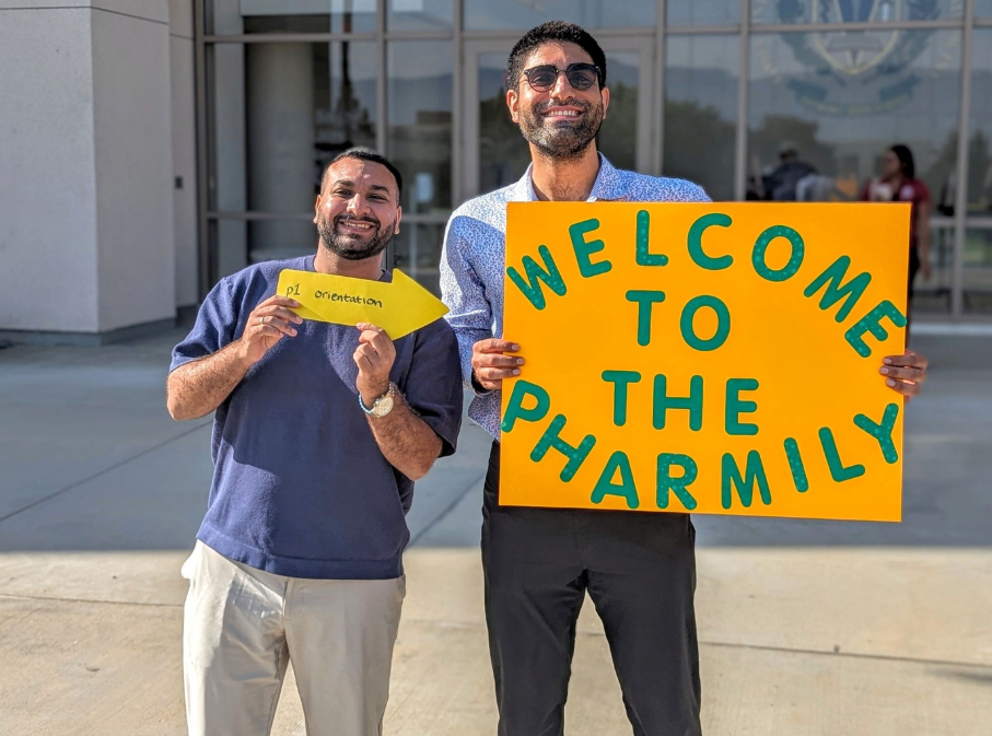 two pharmacy students smiling, standing outside with signs that read "welcome to the pharmily" and "this way to P1 orientation"